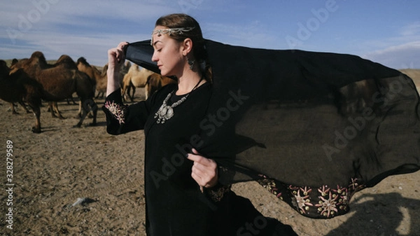 Obraz beautiful woman national eastern clothes black color,veil,hijab,abaya ornament her head,portrait steppes camels,strong wind,desert,wildlife,well,decoration,caucasian,slow ,sun