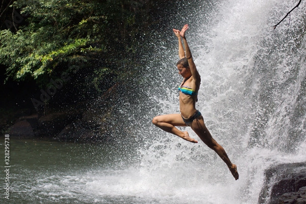 Fototapeta Young athletic woman jumping into water with raised hands. Background of many drops of waterfall and tropical forest