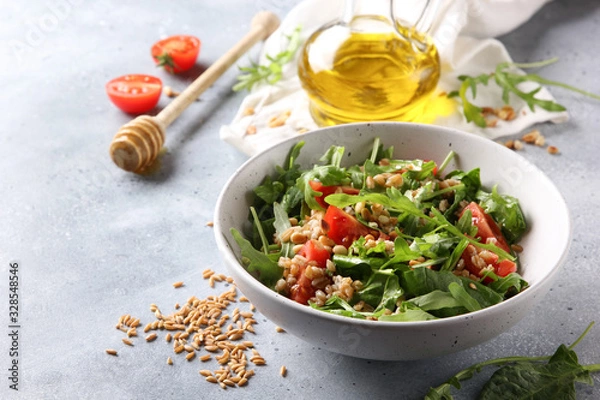Fototapeta The concept of healthy food. Leaf salad with grits and pine nuts in a deep bowl. Arugula, spinach, tomatoes, spelt, olive oil, honey on a light background. Background image, copy space