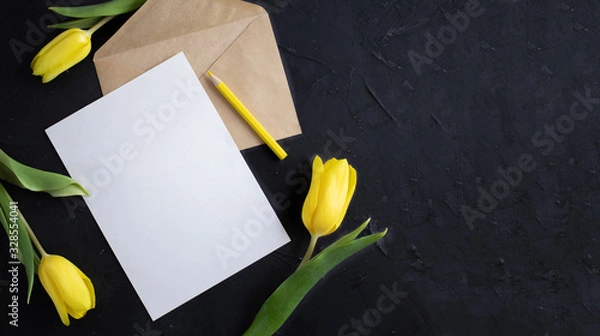 Fototapeta Yellow tulips on the edge of white paper for text on a dark background. A blank sheet of paper with an envelope. Copy space