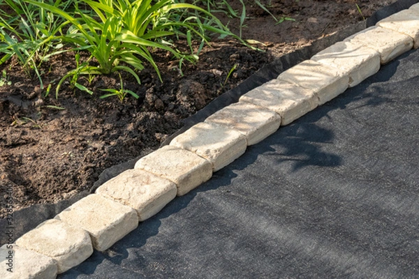 Fototapeta Edging of laid woven geotextile fabric with sandstone pavers in the summer garden under reconstruction
