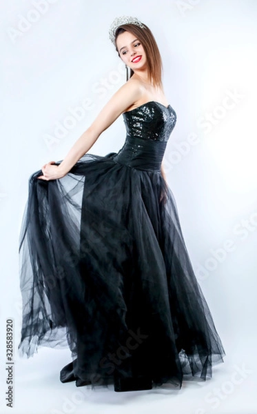 Fototapeta The girl in a black puffy long and diamond diadem is dancing in the studio. Holds a long skirt with his hands. Isolated