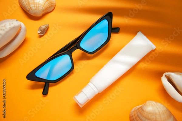 Fototapeta Fashionable glasses and a tube of moisturizer or sunscreen on a yellow background with sea shells and shade from a palm leaf. The concept of self-care , sun protection and safe rest.