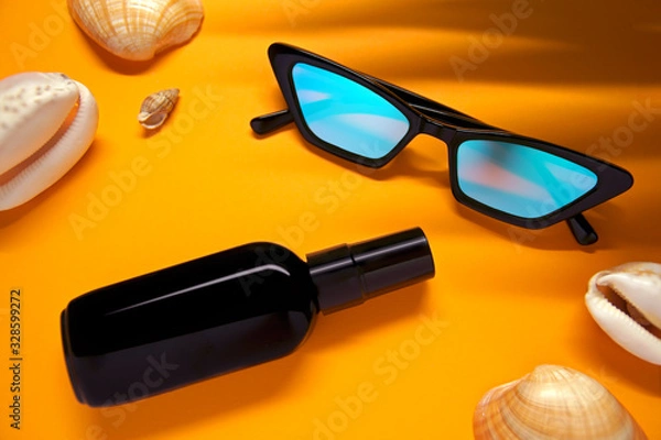 Fototapeta Top view of fashionable glasses and a bottle of moisturizer or sunscreen on a yellow background with seashells and shade from a palm leaf.