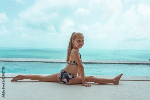 Obraz A small and beautiful girl with red hair and freckles sits by the pool in a bathing suit in the villa and poses in front of the camera. Children's fashion