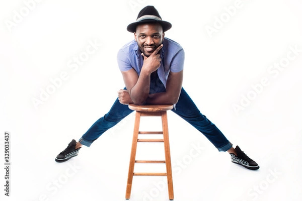 Fototapeta Urban African man having fun during the photo session in the studio. Interior photo of a tall, smiling black man with a hat on a white background