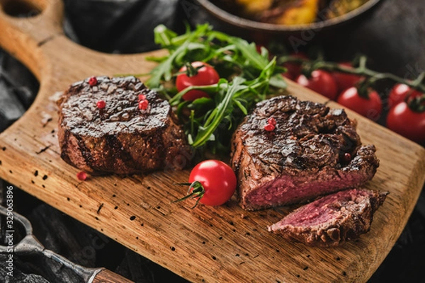 Obraz Grilled fillet steaks on wooden cutting board. Succulent thick juicy portions of grilled fillet steak served with tomatoes and roast potatoes on an old wooden board.