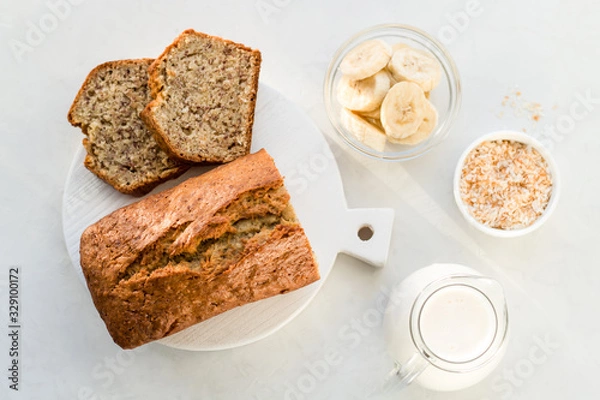 Fototapeta Banana bread with coconut and milk on a light gray background. Homemade delicious pastries. Top view, close- up, copy space.