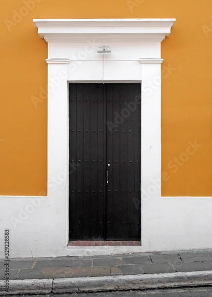 Fototapeta Weathered wooden doors set into an orange wall with white wooden trim along cobblestone sidewalk in old town San Juan.