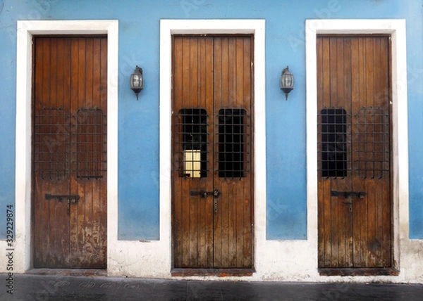 Obraz Three old weathered wooden doors with wrought iron covered widows framed into a vibrant blue wall and surrounded by weathered white trim on a black brick sidewalk.