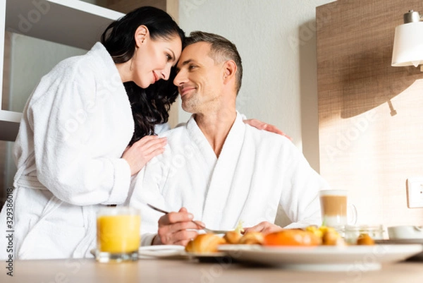 Fototapeta selective focus of smiling girlfriend in bathrobe hugging boyfriend during breakfast
