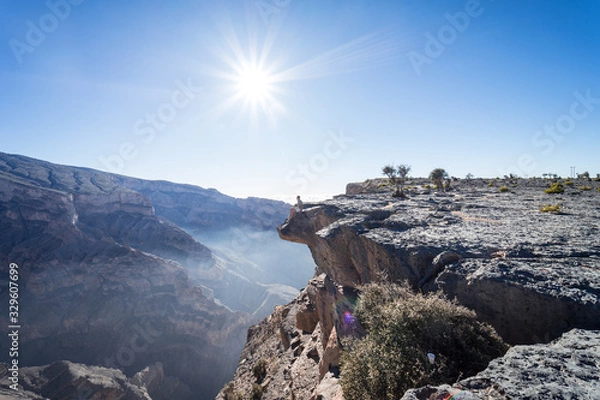 Fototapeta Man peeking over the cliff with the sun in the background at the Grand Canyon of Jebel Shams, Oman