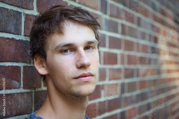 Fototapeta Portrait of a young man leaning against a brick wall with a skeptic and serious expression on his face.