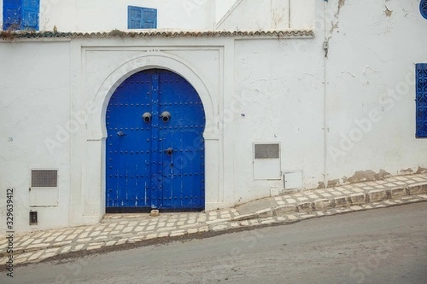 Fototapeta Blue doors in a white wall, the entrance to the house on a sloping street in the city of Sidi bou Said