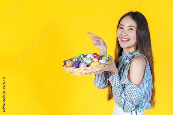 Fototapeta Asian young woman pointing finger to blank copy space and looking at camera, hold basket filled easter eggs, toothy smiling happy face. Holidays and people concept isolated on yellow color background