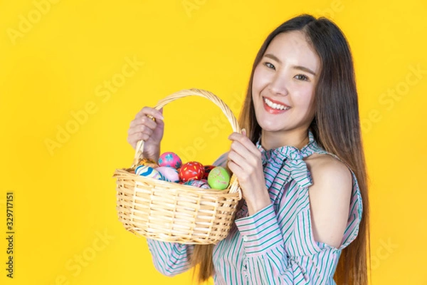 Fototapeta Attractive young Asian woman standing, looking at camera, hold basket filled easter eggs, toothy smiling happy face. Holidays and people concept isolated on blank copy space, yellow color background.