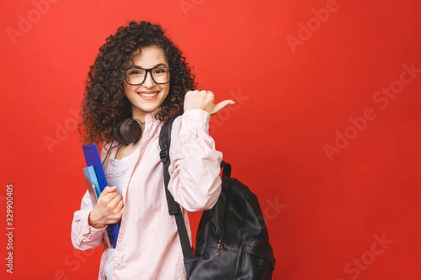 Fototapeta Young curly student woman wearing backpack glasses holding books and tablet over isolated red background. Pointing finger.