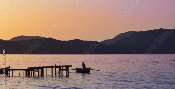 Fototapeta Golden sunset colorsover serene water near Corfu, with a fisherman rowing a boat near a timber jettyy