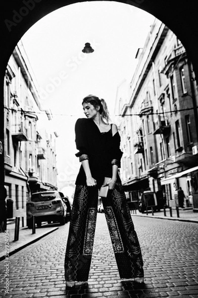 Fototapeta An attractive European girl posing in a street. She dressed in a dark clothes. Street photography. Black and white photo. Soft focus.