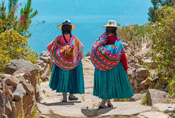 Fototapeta Dwie tubylcze kobiety Quechua w tradycyjnych strojach idące ścieżką do portu Isla Taquile (wyspa Taquile) z jeziorem Titicaca w tle, Peru.
