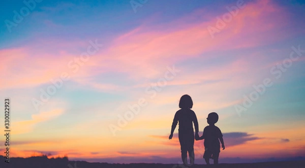 Obraz Silhouette back view of two children sibling walking on the beach while holding hands with sunset background.A silhouette of big sister and little brother looking GOD creation beautiful sunset sky.