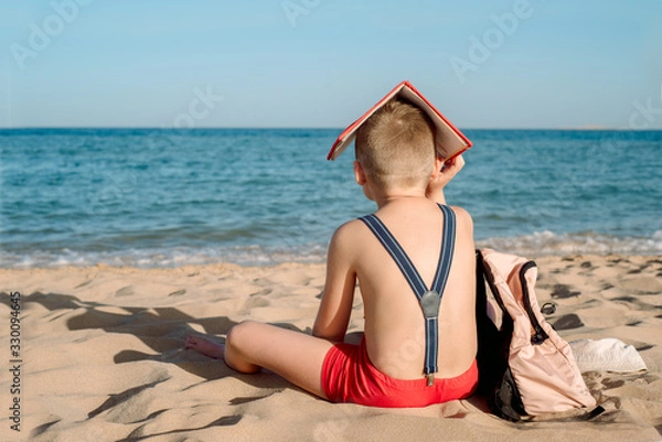 Obraz Happy young boy reading a book on a sunny dayl. boy sitting with his back at the beach. Last day of vacation. Back to school. Backpack and suspenders, red book. Bad mood and not a desire to learn.