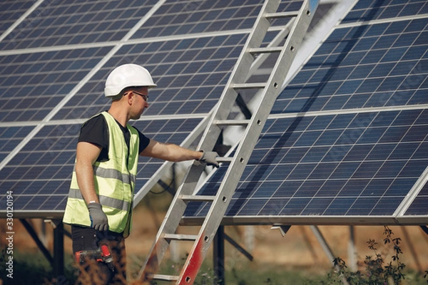 Fototapeta Engineer in a white helmet. Man near solar panel. Worker with a ladder