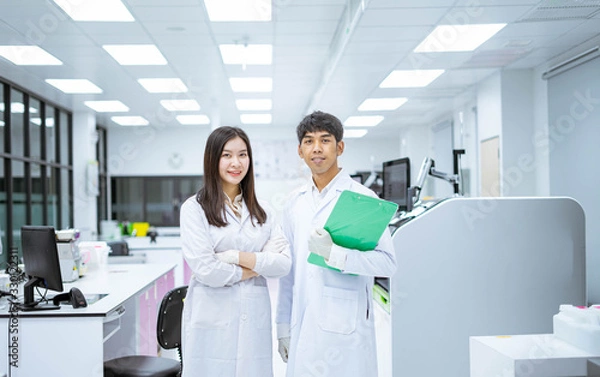 Fototapeta two young smiling scientist in white lab coat standing with automation blood analyzer at medical laboratory 