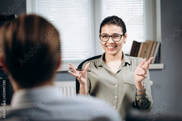 Fototapeta Portrait of smiling beautiful lady in stylish blouse and eyeglasses helping male patient to deal with emotional excitements. Concept of professional consultation and psychology