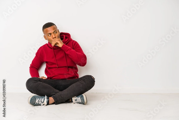 Fototapeta Young latin man sitting on the floor isolated thoughtful looking to a copy space covering mouth with hand.