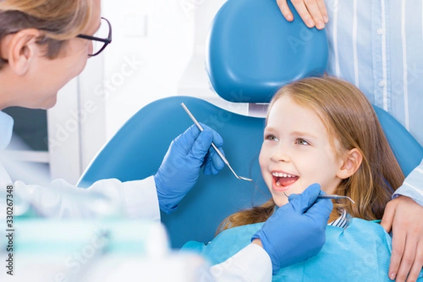 Fototapeta Little cute smiling girl is sitting in dental chair and opening mouth in clinic, office. Doctor is preparing for examination of child teeth with tools, instruments. Visiting dentist with children.