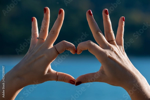 Obraz Female hands in heart shape in focus, blue sea and beachfront on background 