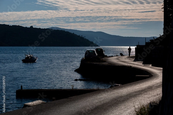 Obraz Silhouette of a person walking on beachfront asphalt road, silhouettes of cars on the left side of the road,  partly cloudy sky, contour light