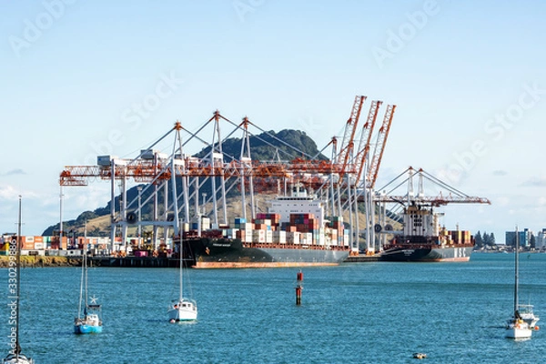 Fototapeta TAURANGA, NEW ZEALAND - MARCH 6, 2020: Cargo ships docked into Tauranga Harbour Port waiting for the adjacent container cranes to load. Mount Maunganui in the background.