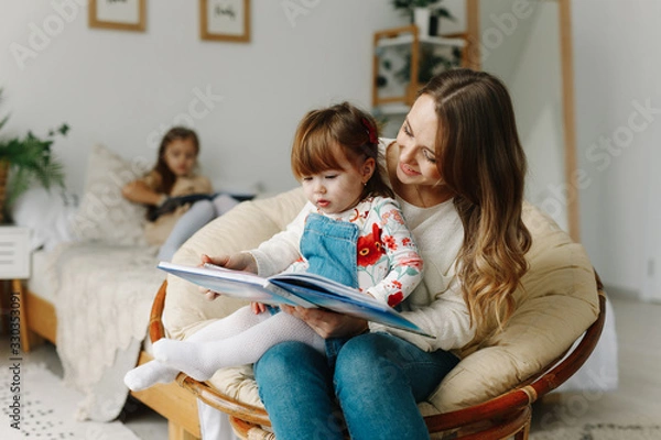 Obraz Portrait of a smiling young cute mother and two daughters reading a book relax in the sofa in a bright big white room