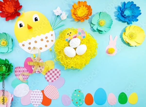 Obraz Child made crafts his own hands at Easter. Handmade nest with eggs, flowers, chicken, chicken in an egg, frame of eggs. Cute art creativity on blue background. Flat lay, top view, close up, copy space