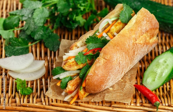 Fototapeta Baguette sandwich with fried chicken and fresh vegetables-carrots, cucumber, daikon, red pepper and cilantro (coriander) on a wicker table. Delicious fast food. Vietnamese Banh Mi. Selective focus