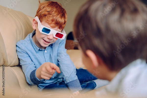 Fototapeta Portrait of a little boy pointing out to his brother an educational three dimension book. Cheerful kid playing with three dimensional glasses and interactive cinema at home. Leisure and movies