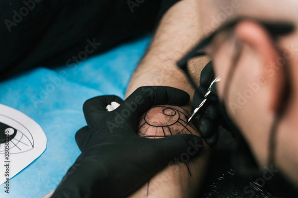 Obraz A selective focus shot of a tattoo artist with black gloves creating a tattoo on a man's arm
