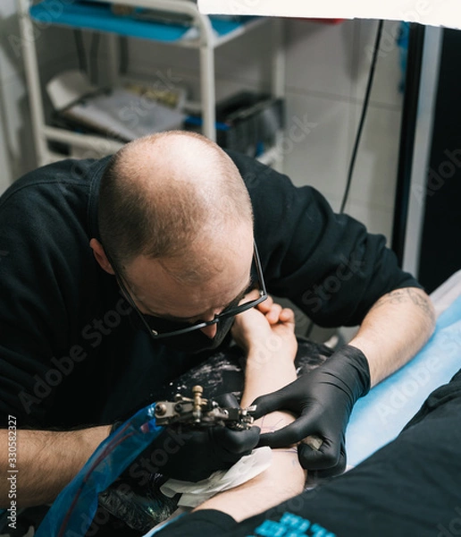 Fototapeta A closeup shot of a tattoo artist with eyeglasses creating a tattoo on a man's arm