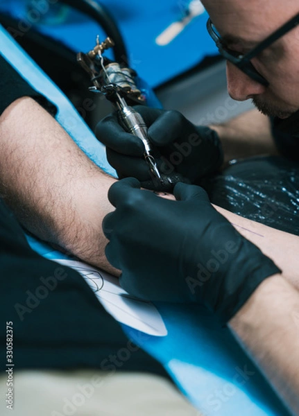 Obraz A closeup shot of a tattoo artist with eyeglasses creating a tattoo on a man's arm