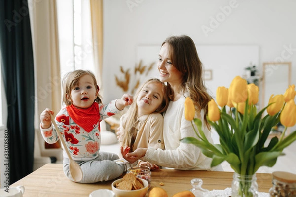Obraz Friendly young family having good time together in a bright kitchen. Little baby girl on the table near tulips playing with a spoon.