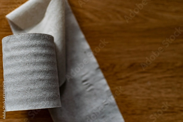 Fototapeta A roll of clean, plain toilet paper, cheap quality, dirty color, lies on a brown wooden table, slightly unwound and with the end torn off