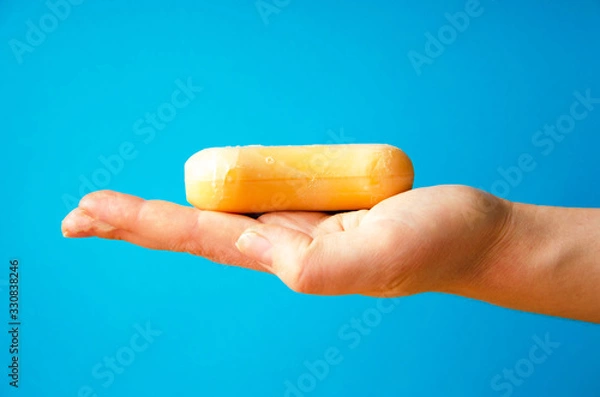 Fototapeta Women's hands are holding soap. Soap foam on the hands. Yellow soap in the hands. Woman washes soap with hands side view on a blue background. Virus protection.  COVID-19