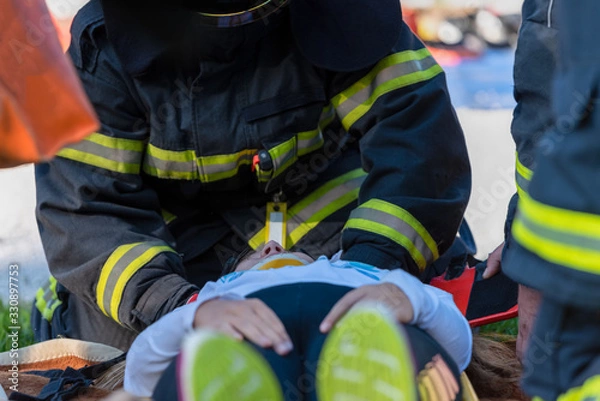 Fototapeta The victim in a car accident lies on the floor of a stretcher. The fireman helps her.