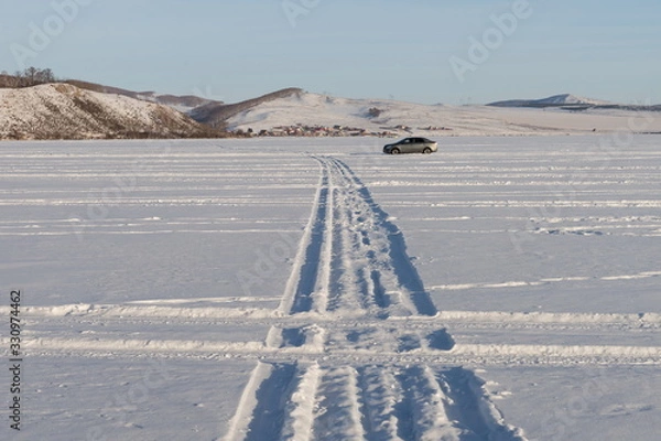 Fototapeta A geometric pattern from the wheels of cars was imprinted on the snowy plain of a lake against the shore.