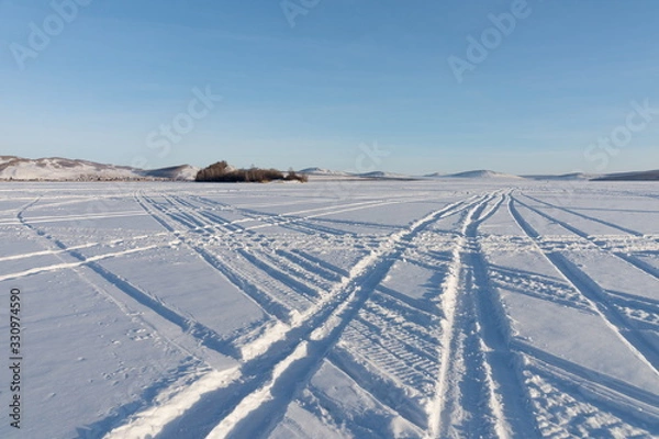 Fototapeta Beautiful geometric patterns from the wheels of cars were imprinted on the snowy plain of the lake against the shore.