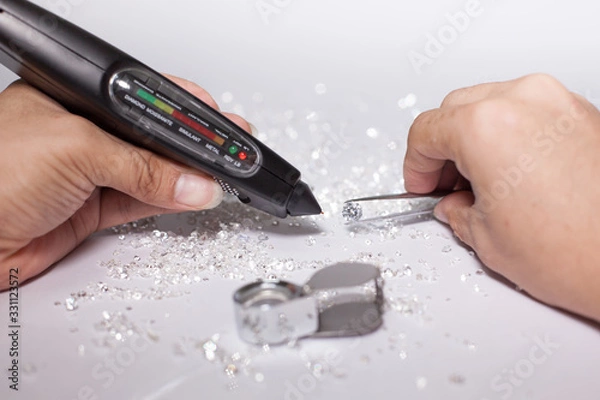 Fototapeta Diamonds on tweezers with testing machine on a white background, reflections on the ground. brilliant cut diamond held by right hand, tester, magnifier , diamond checking equipment.
