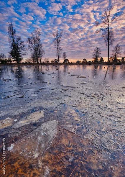 Fototapeta A cold sunset in winter on a frozen lake with a beautiful sky and sunbeams in the background Friesland, The Netherlands
