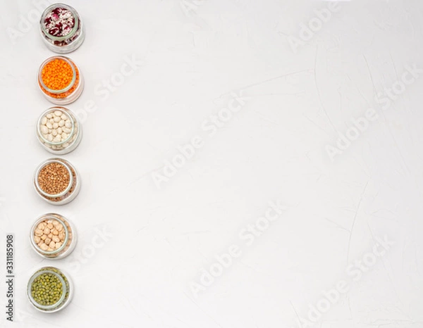 Fototapeta A variety of dry cereals for vegetarians in glass jars: lentils, chickpeas, beans, buckwheat on a white background